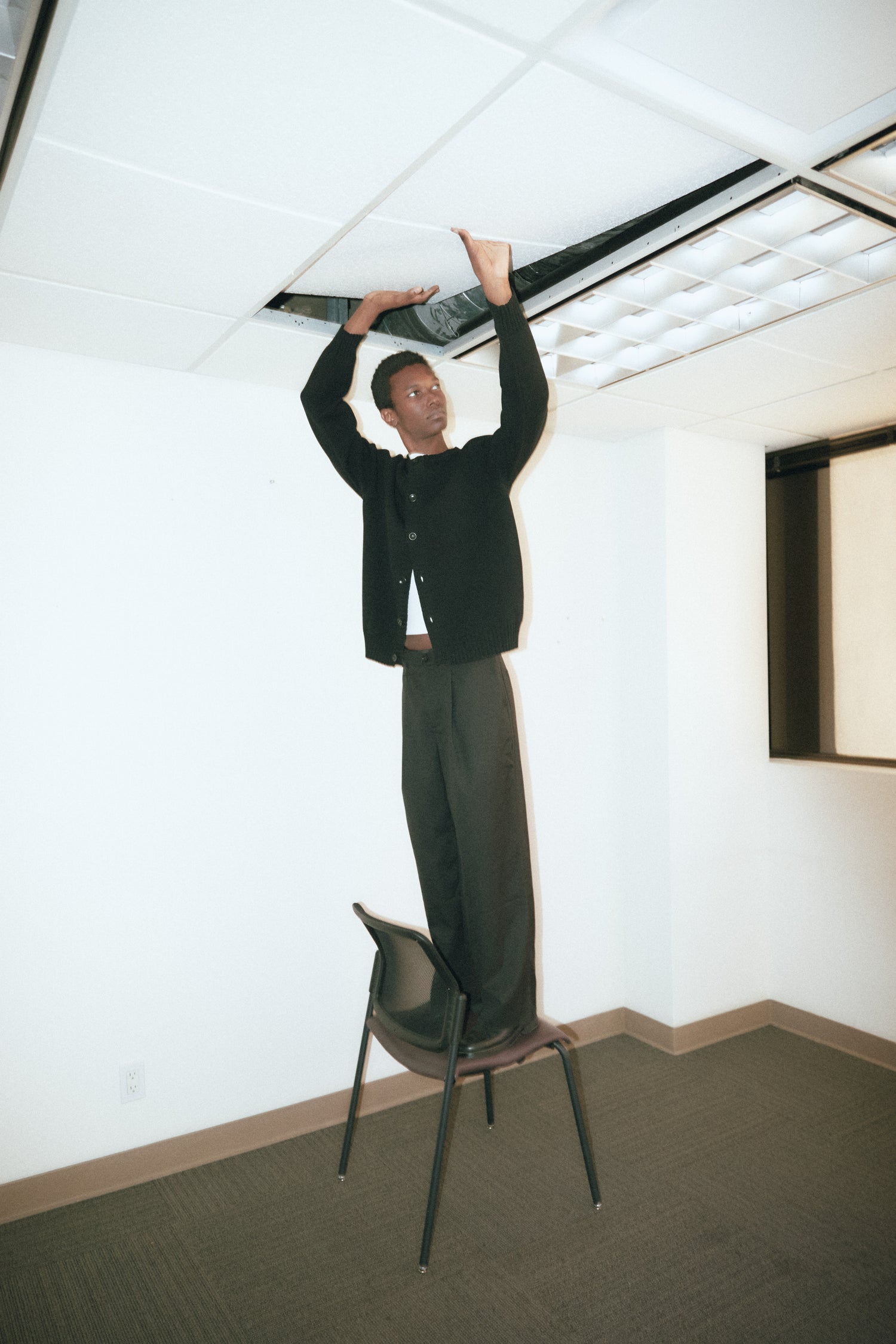 	A man standing on a chair, stretching his arms upward toward the ceiling, wearing a black sweater and wide-leg trousers.