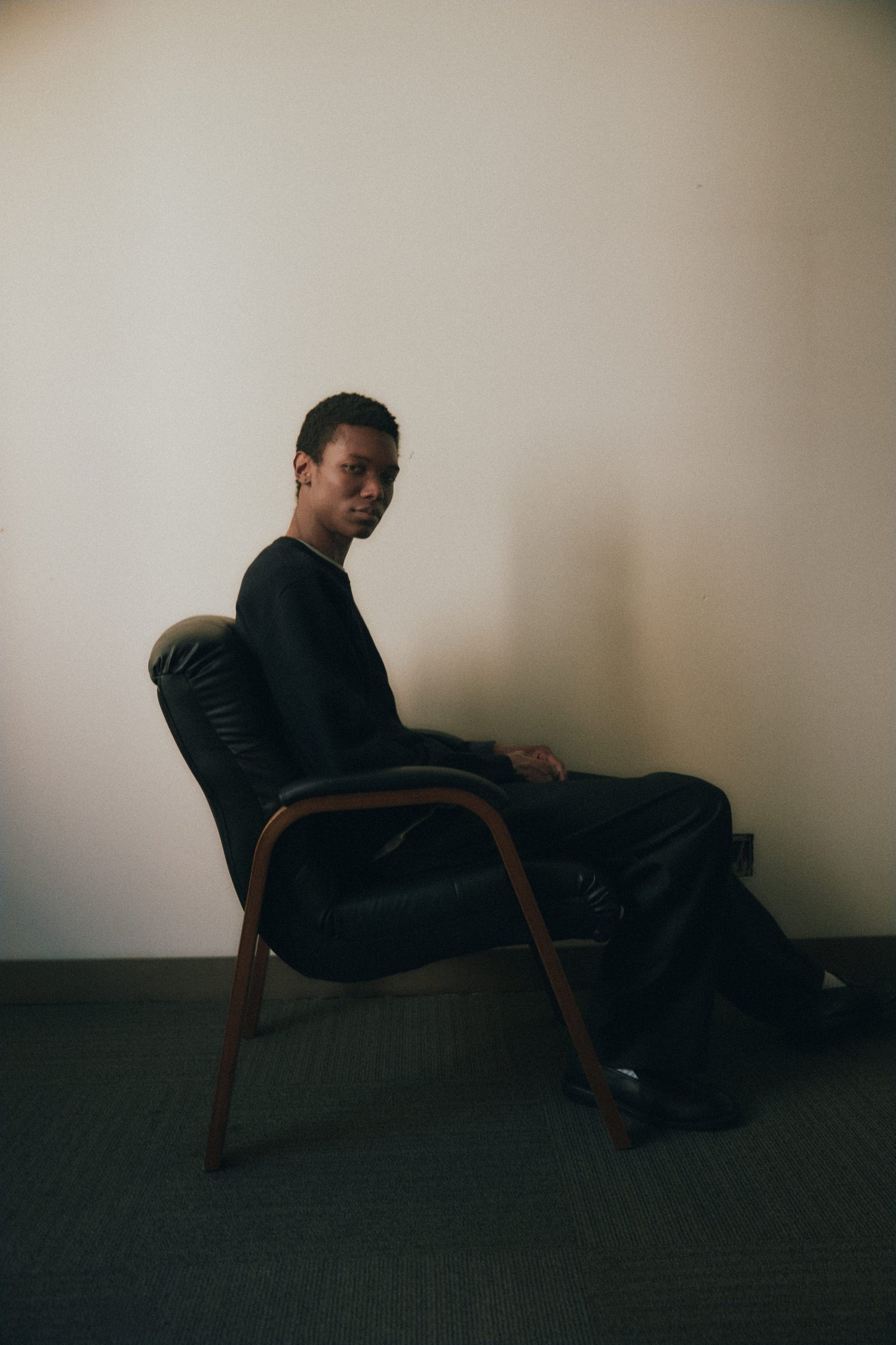 	A man seated in a wooden chair with black leather upholstery, facing sideways, wearing a black outfit in a neutral-toned room.