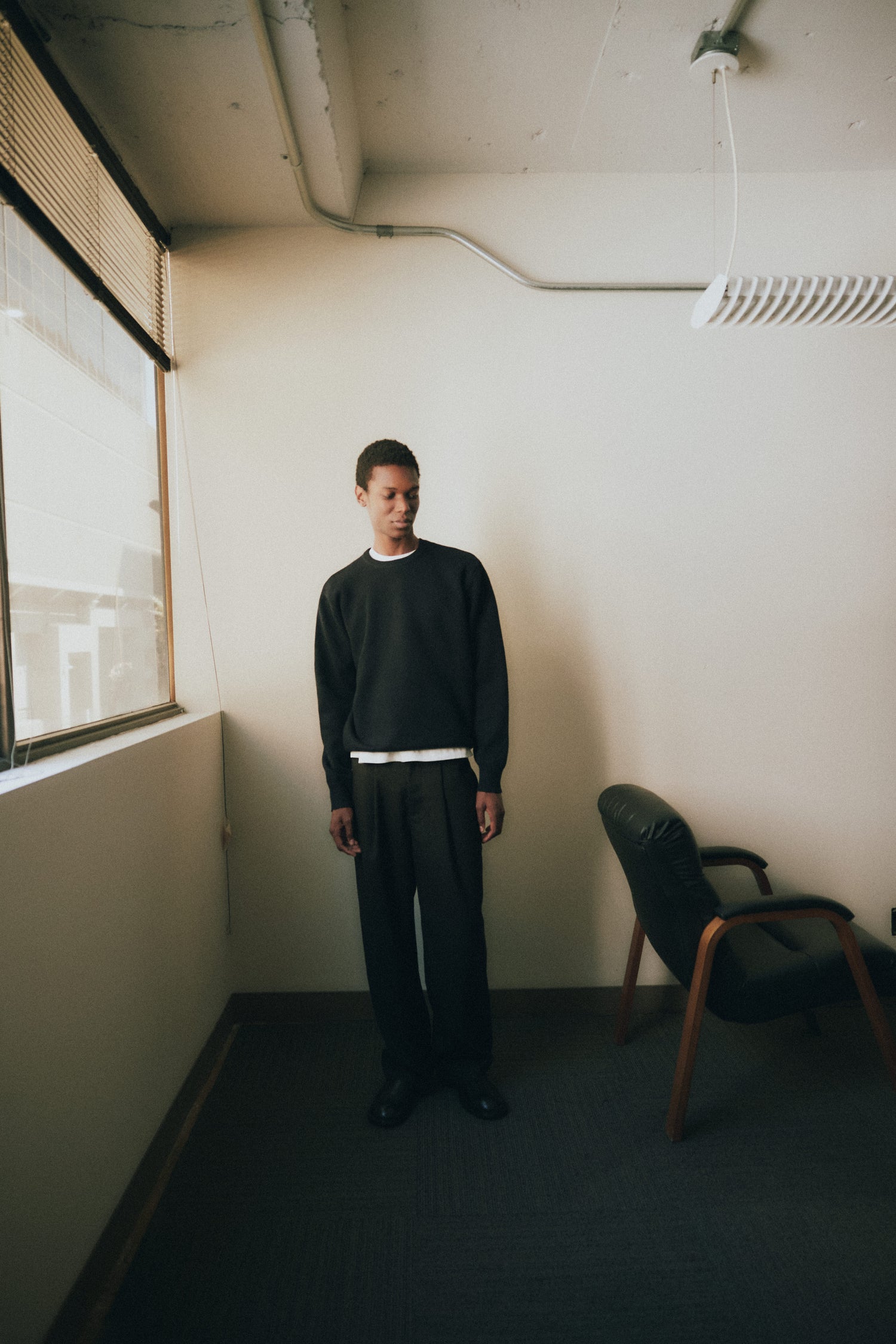A man standing in a minimalist room near a window, wearing a black sweatshirt and loose black trousers, looking slightly to the side.