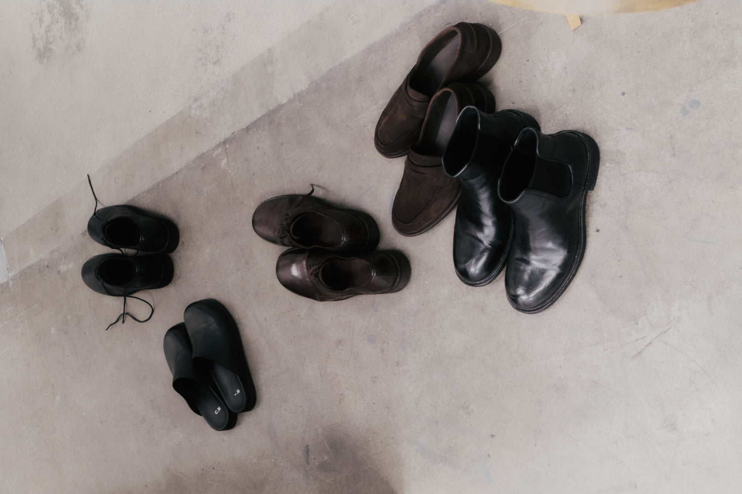 A collection of black and brown leather shoes and sandals placed casually on a concrete floor.