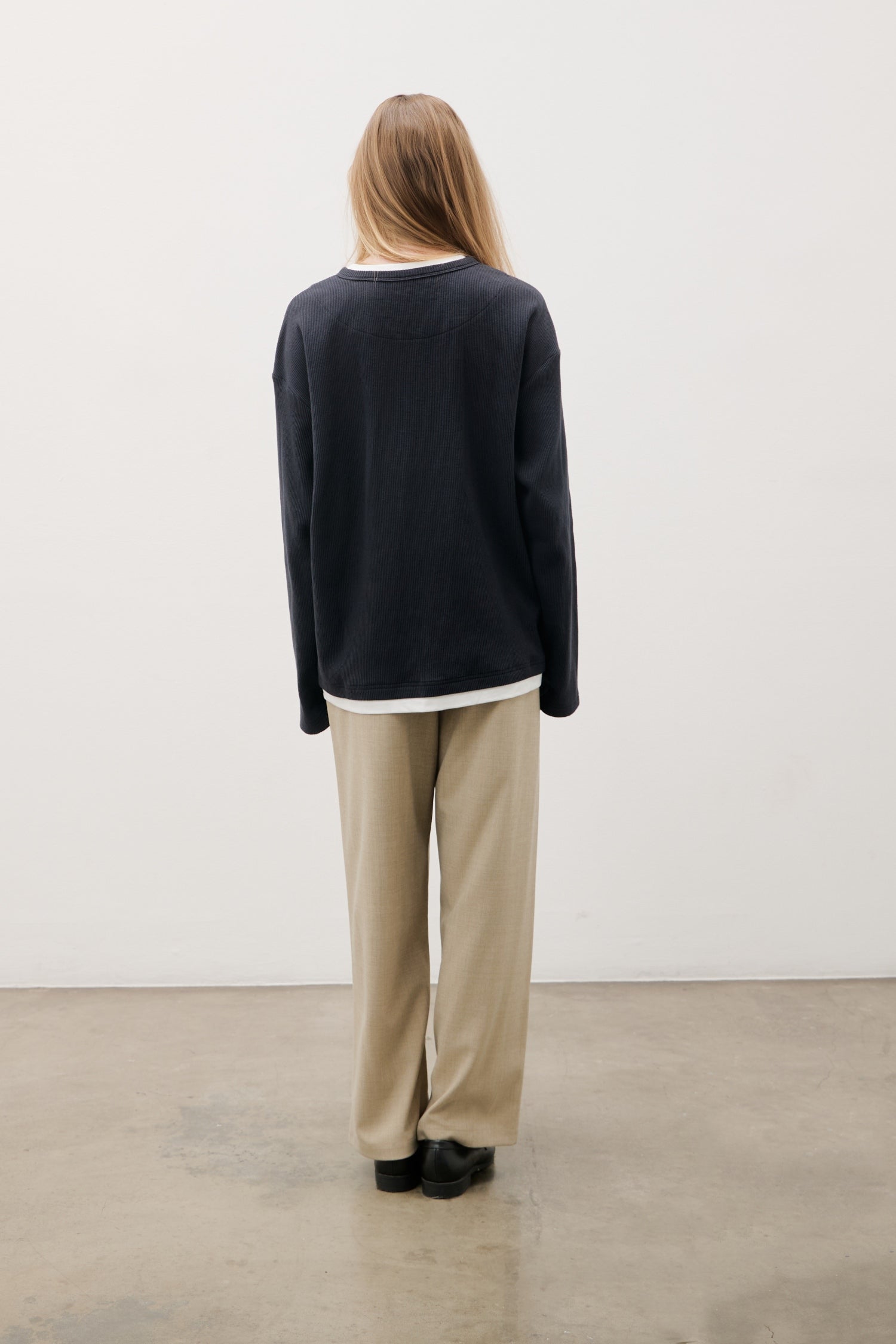 Person wearing a black sweater and beige pants standing against a white wall.