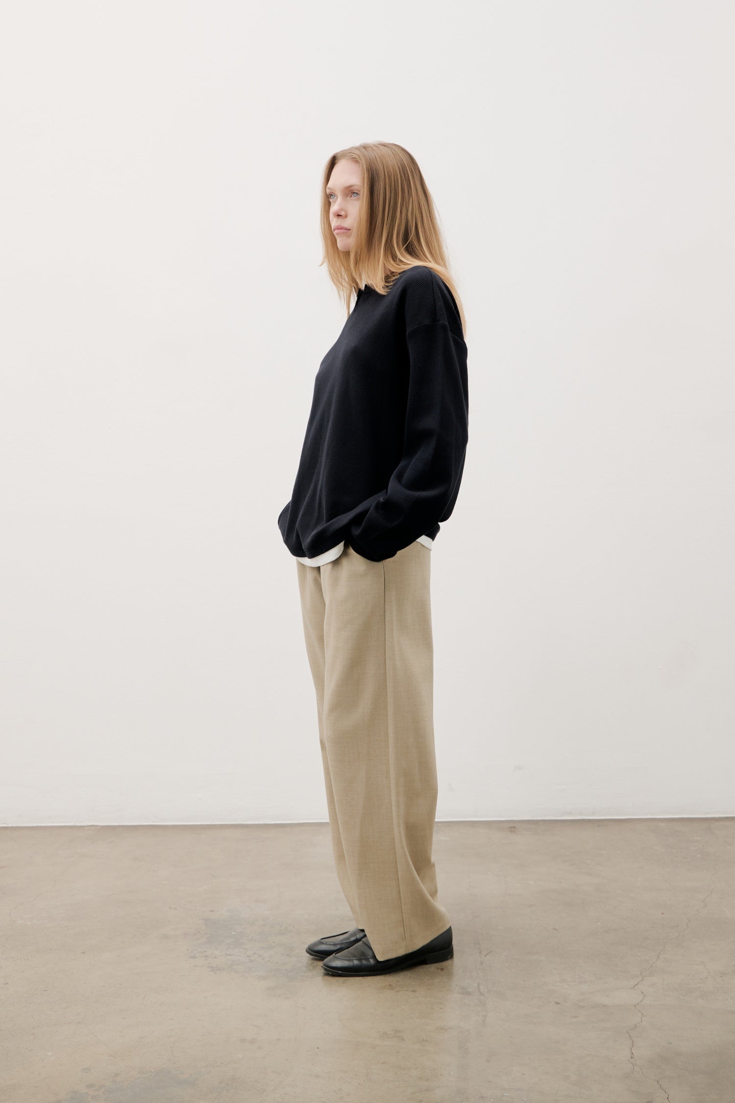 Person wearing a black sweater and beige pants standing against a white wall.