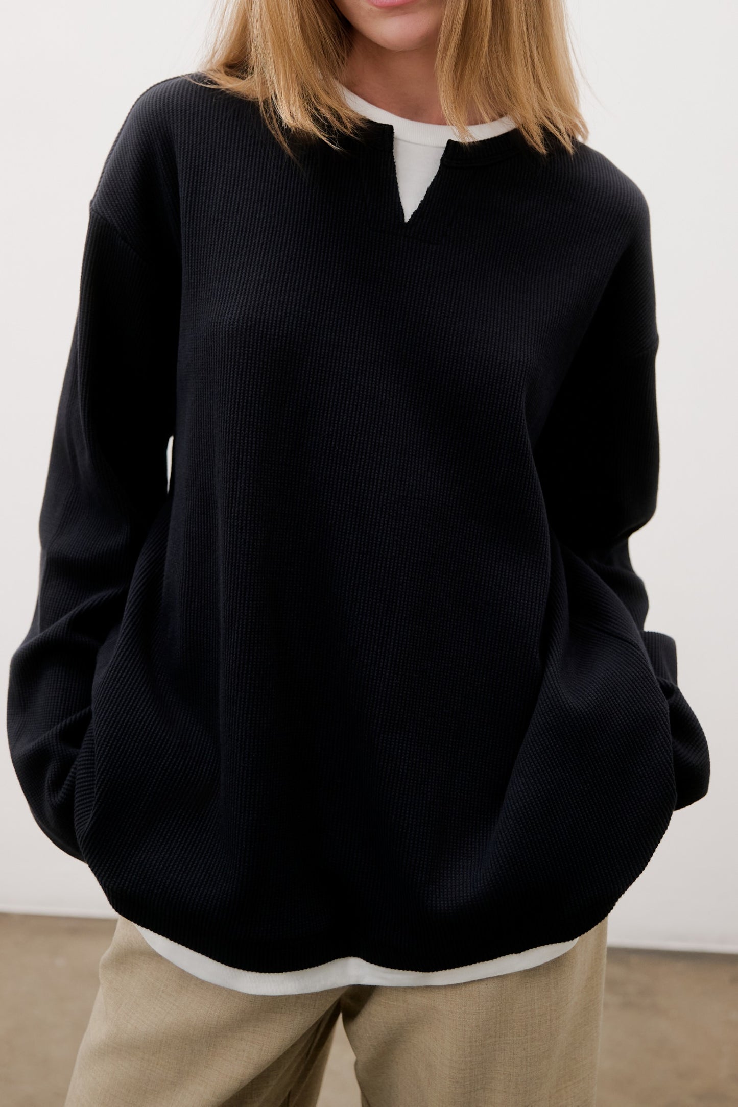 Person wearing a black sweater over a white shirt with beige pants against a plain background