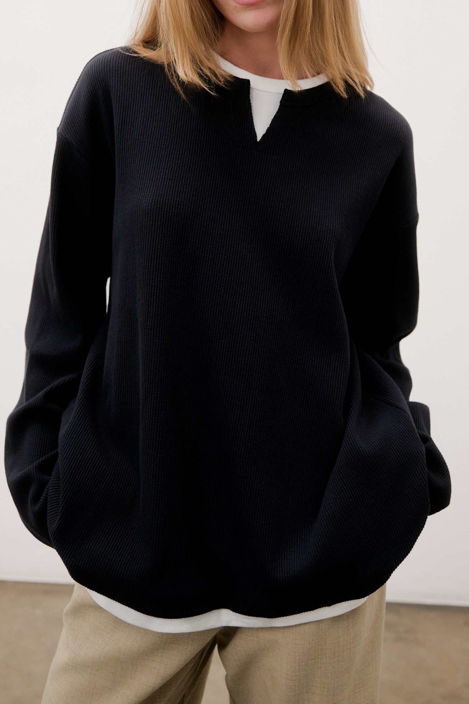 Person wearing a black sweater over a white shirt with beige pants against a plain background