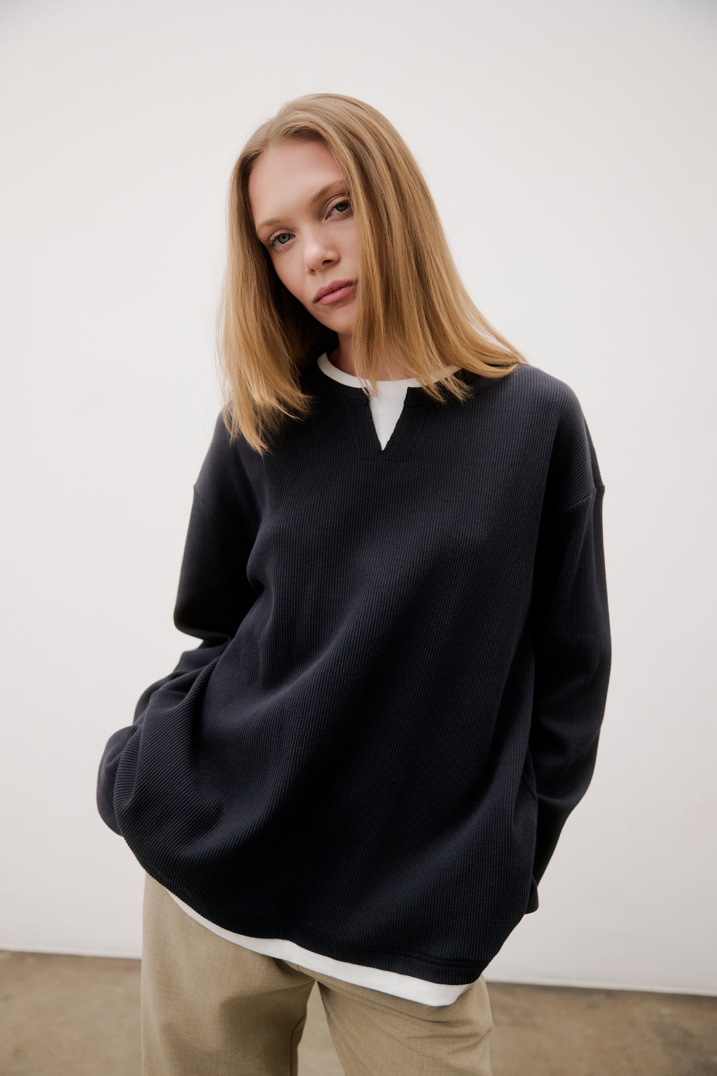 Person wearing a black sweater over a white shirt with beige pants against a white background
