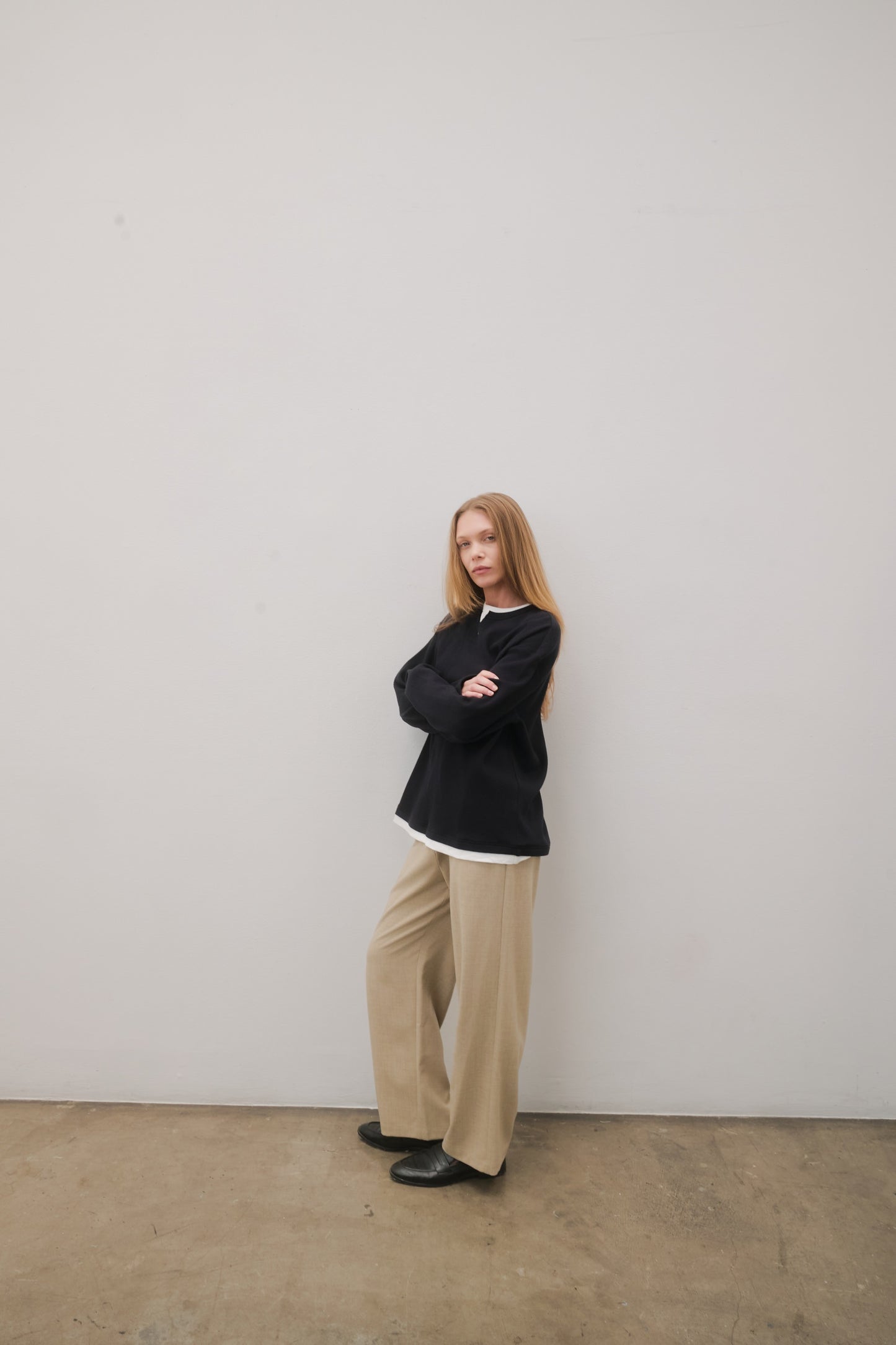 Person wearing a black sweater and beige pants standing against a plain white wall.